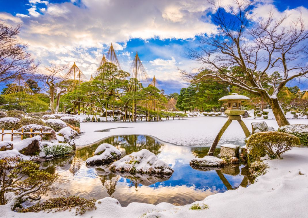 Giardino giapponese coperto di neve con laghetto, lanterna di pietra e alberi protetti da corde sotto un cielo invernale.