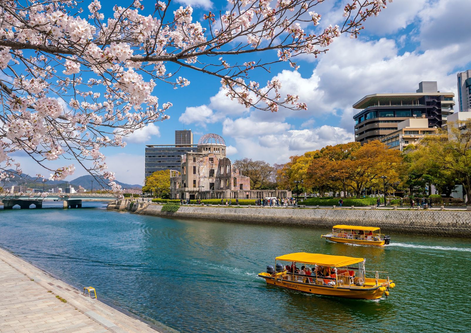 Barche che navigano lungo il fiume Motoyasu a Hiroshima in una giornata di sole con i fiori di ciliegio davanti